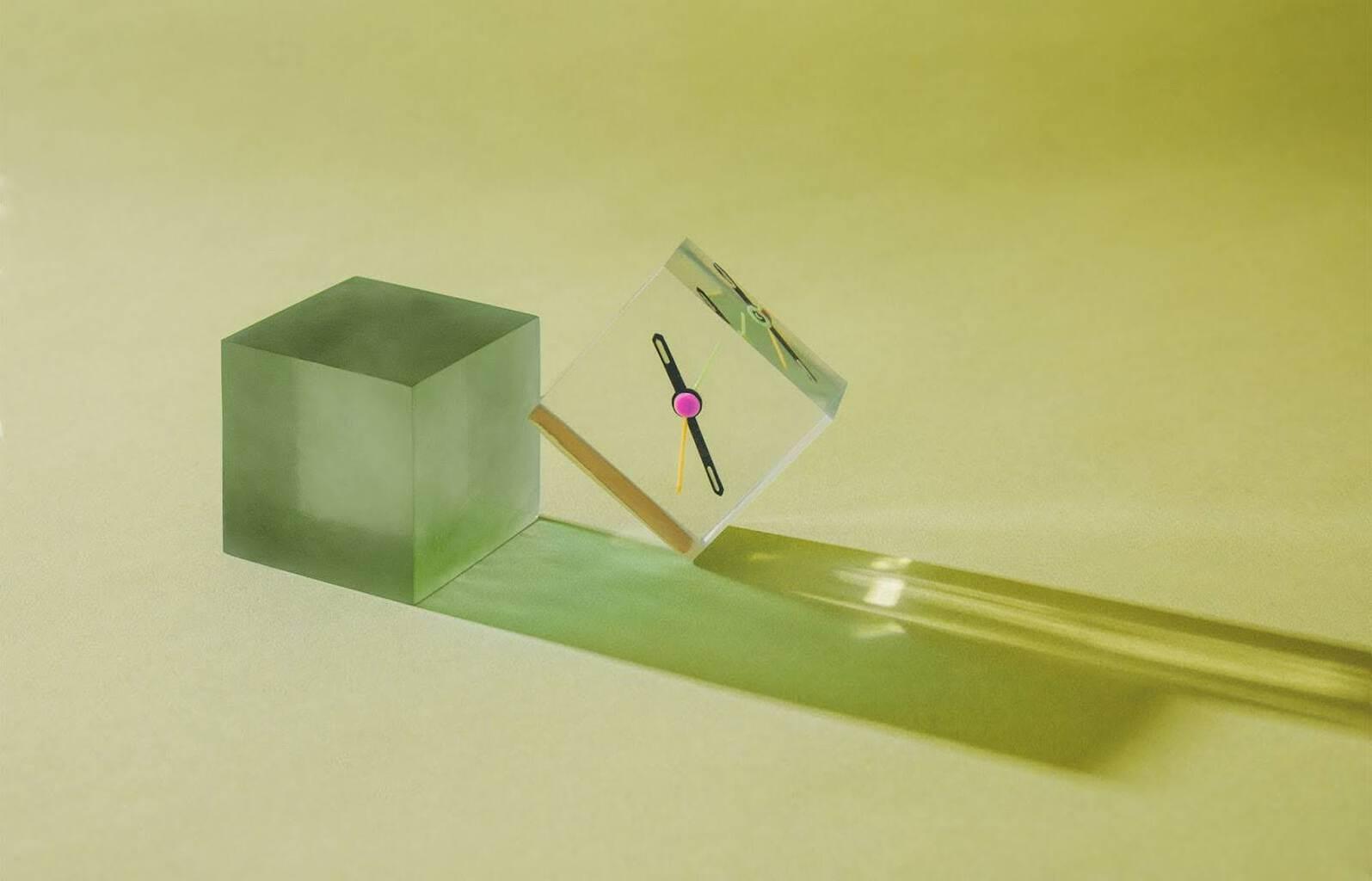 A clear cube with clock hands inside sits next to an opaque green cube.