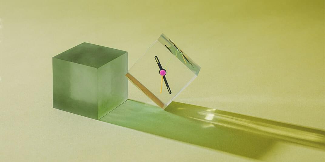 Clock hands are fixed to a clear cube that leans against a larger green cube. 