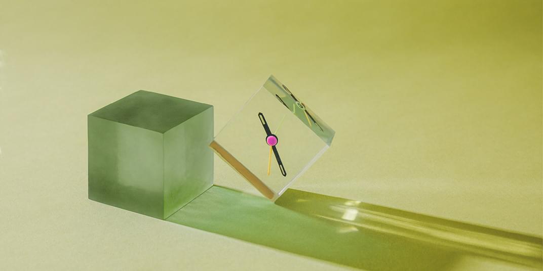 A clear cube with a clock hand in it leans against a green cube, both creating a reflection on the ground.