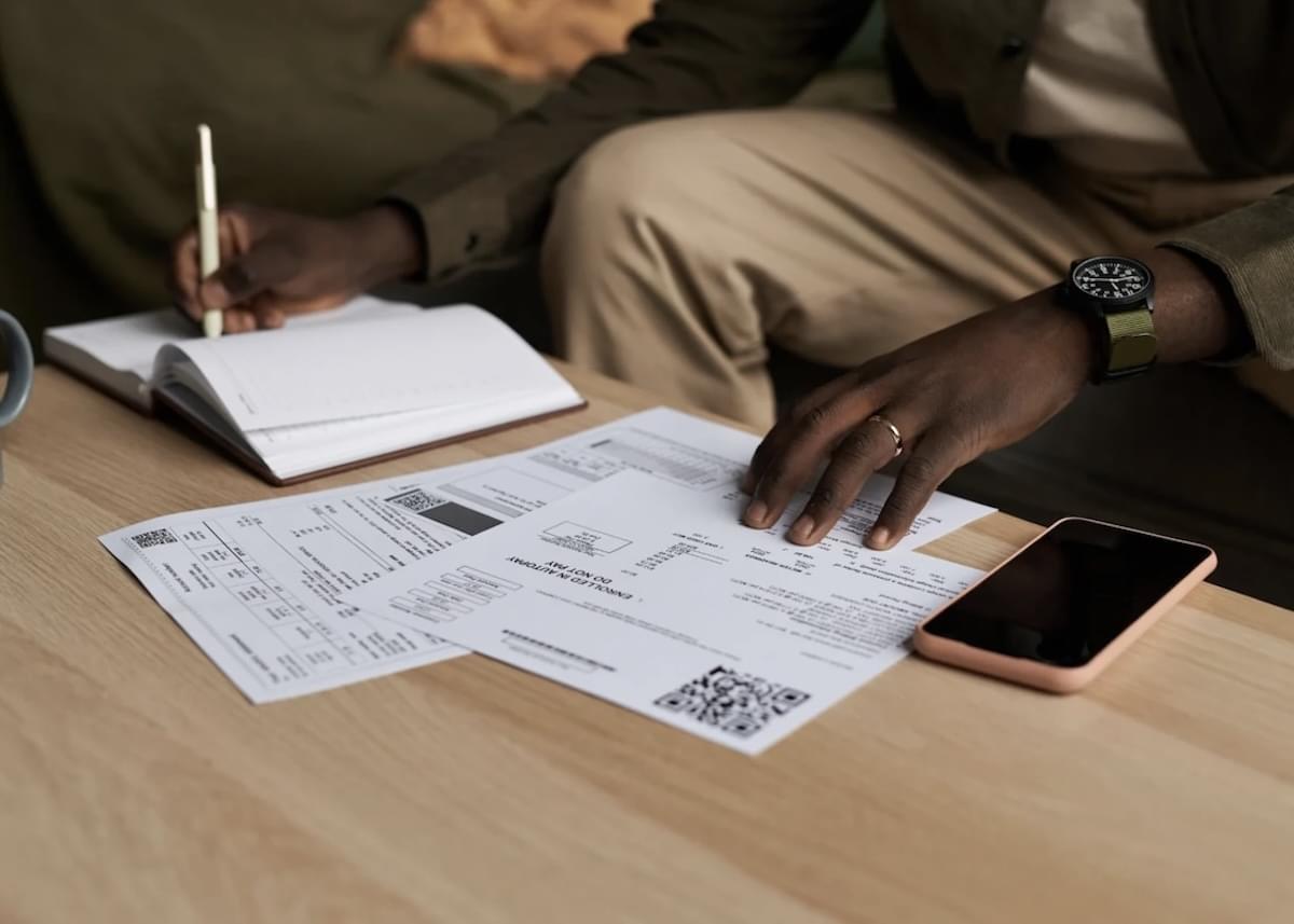 A cropped photo of a man writing in a notebook and working on document next to a mobile phone at a coffee table.