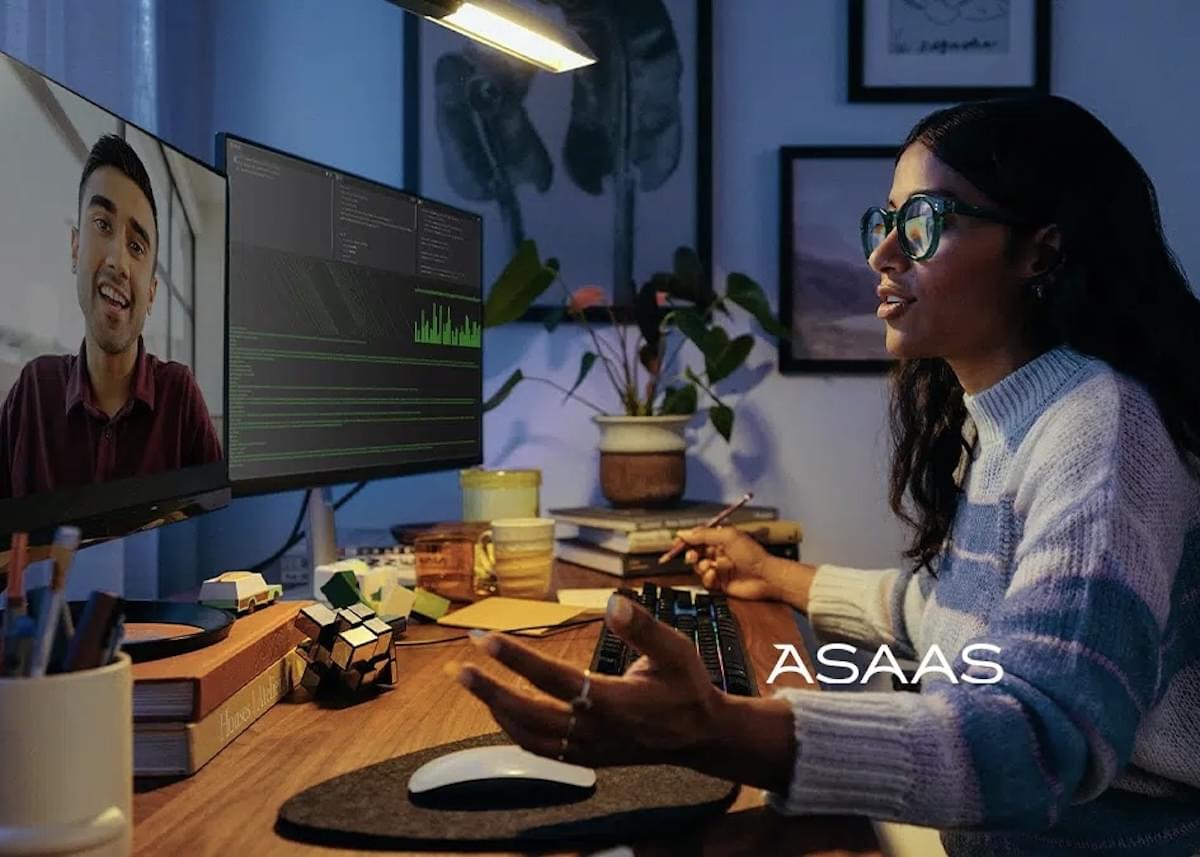 A woman in a dimly lit office talks with a man over a computer screen. A second monitor shows computer code.