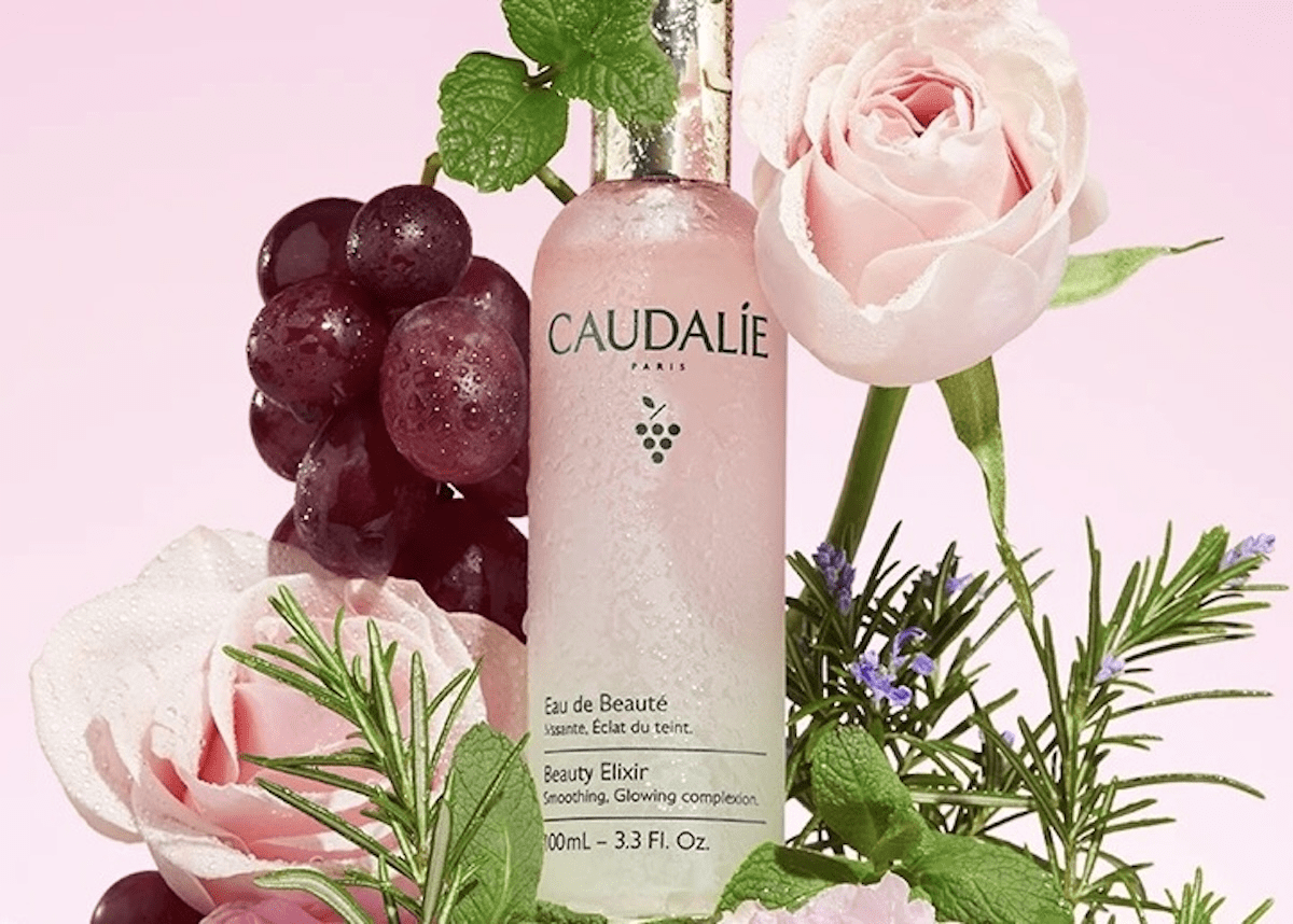 A 3.3 fl. oz. bottle of Caudalie Beauty Elixir surrounded by flowers against a pink background