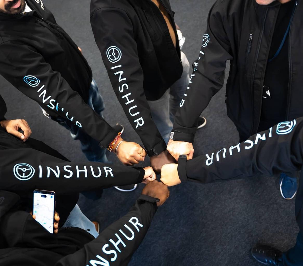 Six INSHUR team members wearing branded black long-sleeved shirts stand in a circle and touch fists