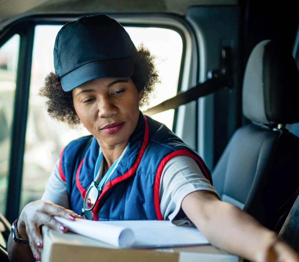 A woman in a delivery uniform sits in a parked van, looking at a package and clipboard with a slight smile