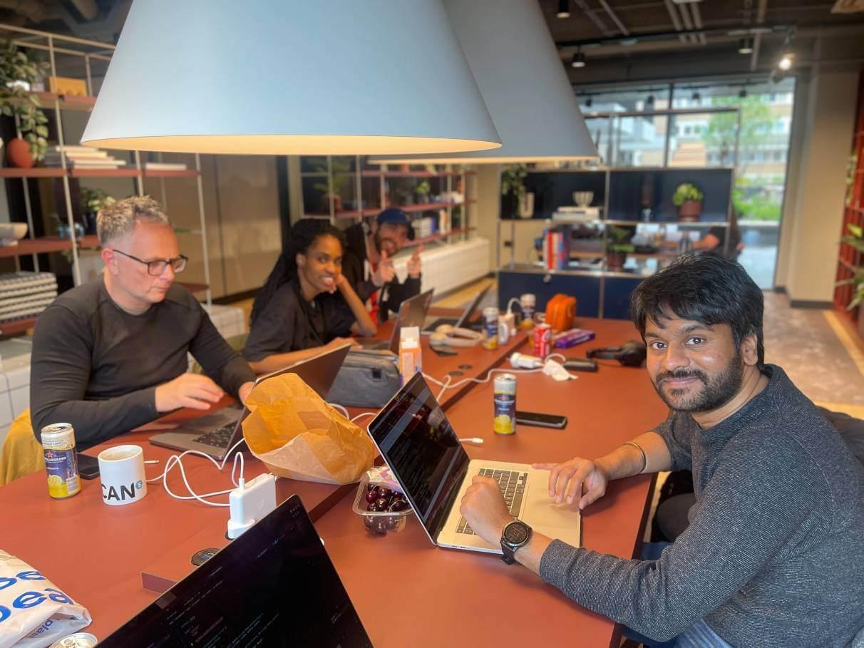 Four people in an open office work on their laptops, with three of the people smiling and looking at the camera