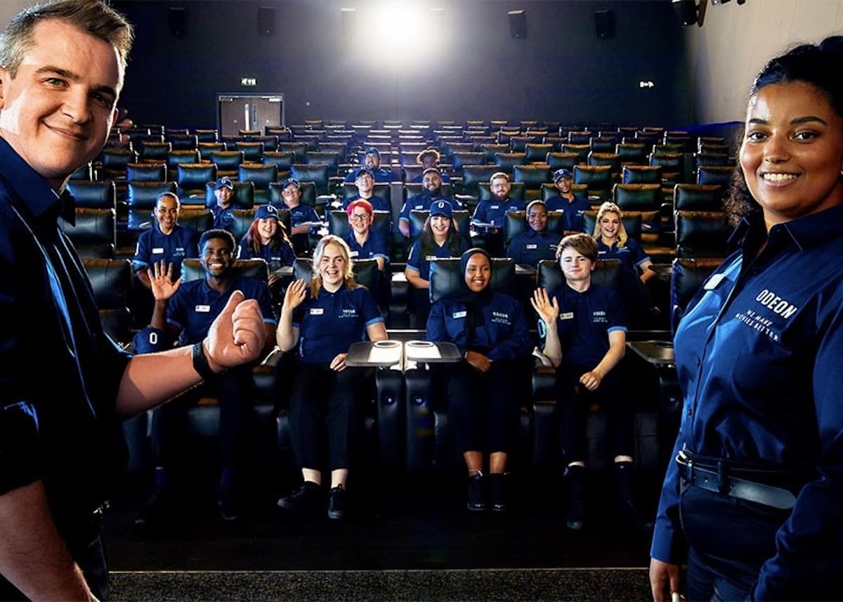 A man and a woman in ODEON uniforms stand in the foreground of employees sitting inside a theater.