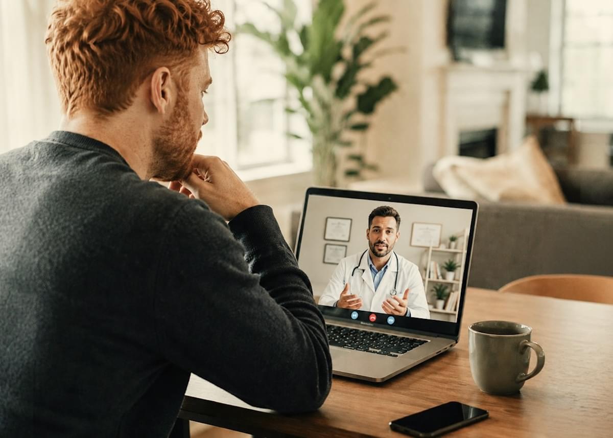 A man sits in light-filled room at a table, with his laptop open, consulting with a man in a labcoat online.
