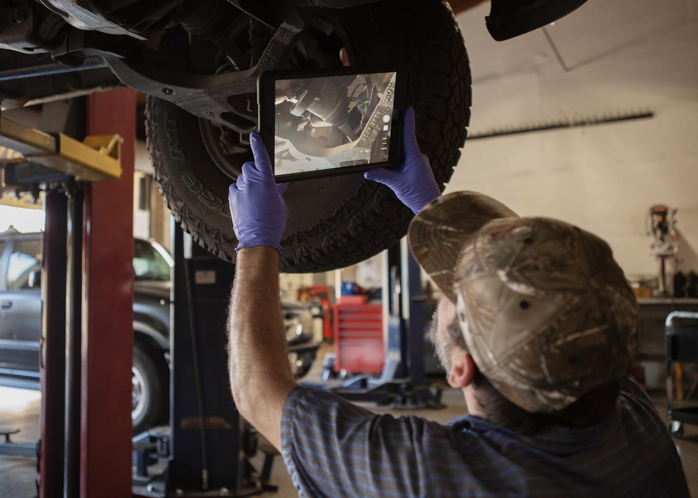 A man in an auto workshop takes a closer look at a tire on a car lift using a tablet.