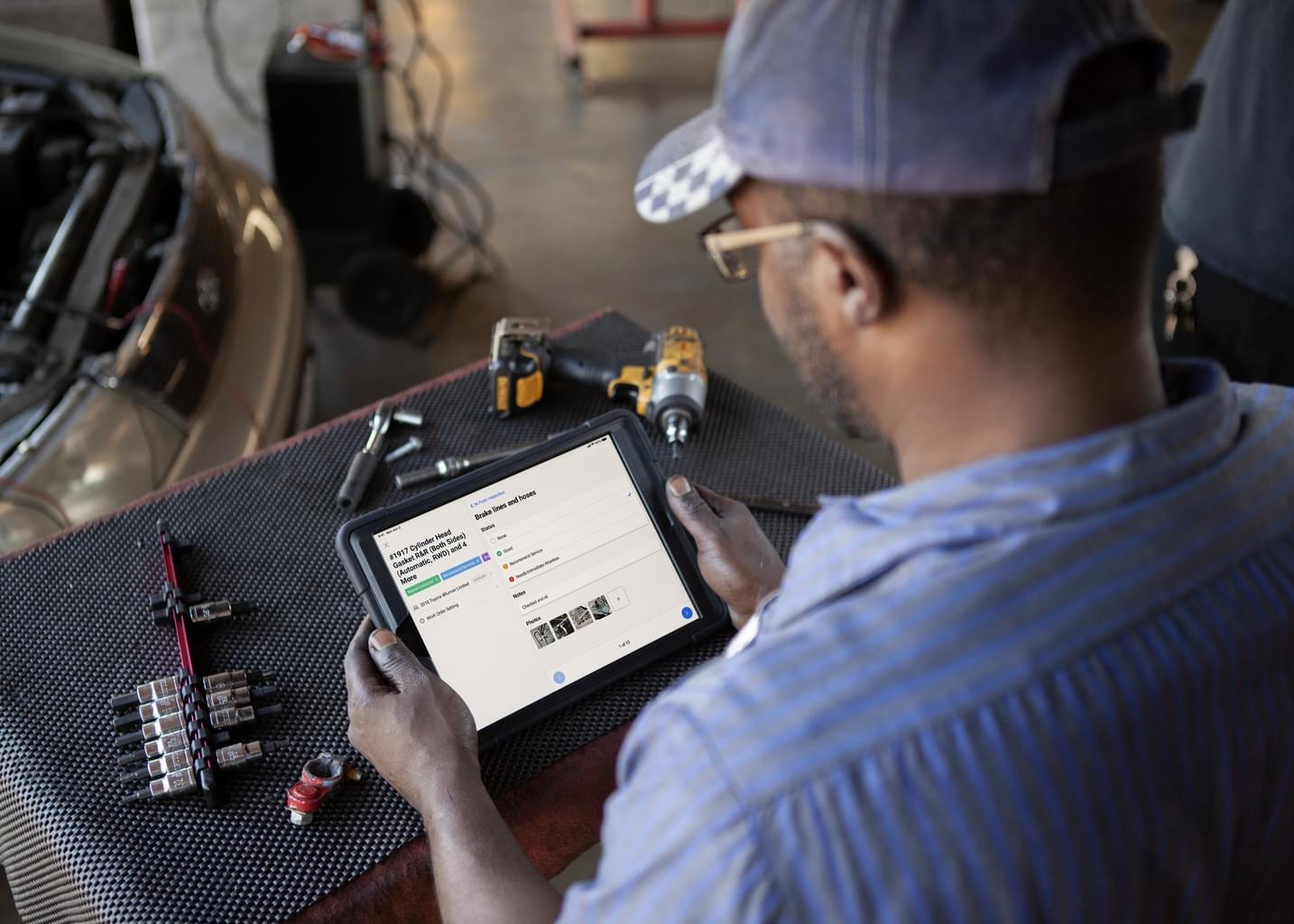 A man wearing a baseball cap in an auto workshop looks at the Shopmonkey website on a tablet.