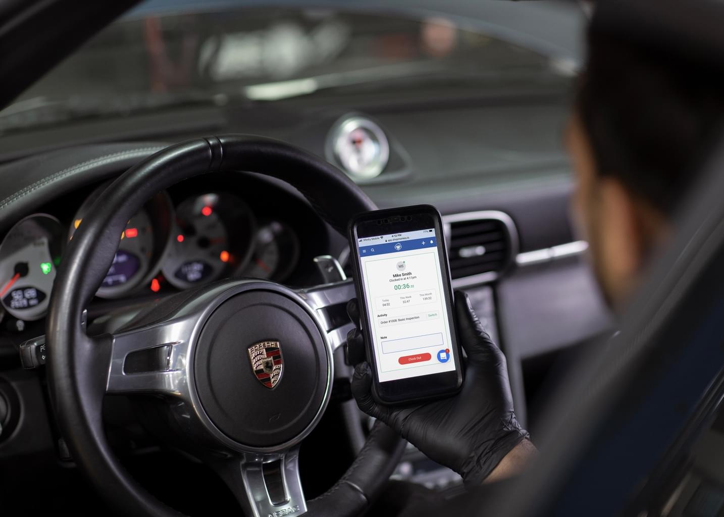 Sitting inside a Porsche, a man wearing a black leather glove looks at the Shopmonkey website on his mobile phone.