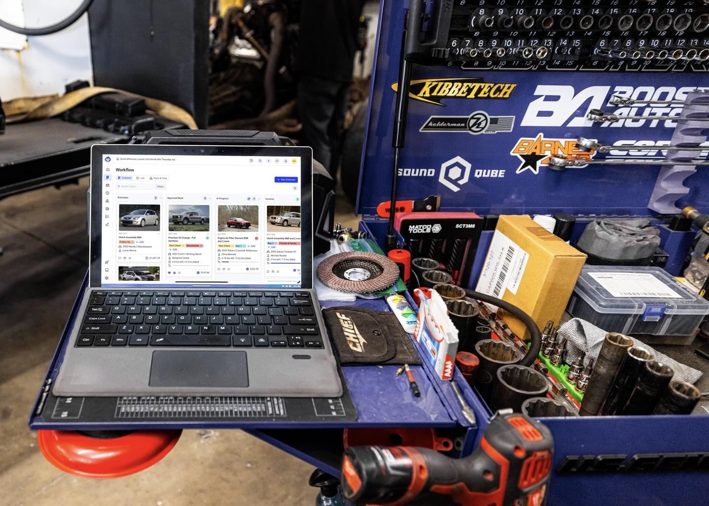 Inside an auto workshop, a laptop sits on a cluttered work table open to the Shopmonkey website.