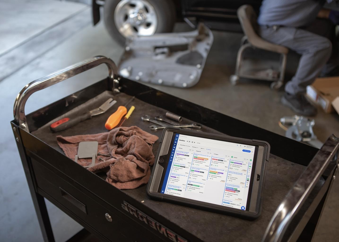 Inside an auto workshop, a tablet sits on a cluttered work table open to the Shopmonkey website.