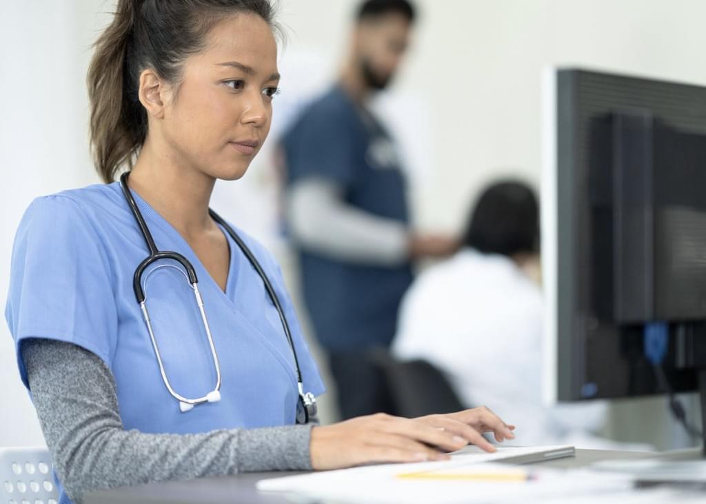 A woman wearing light blue scrubs with a stethoscope around her neck, stands as she types on a computer