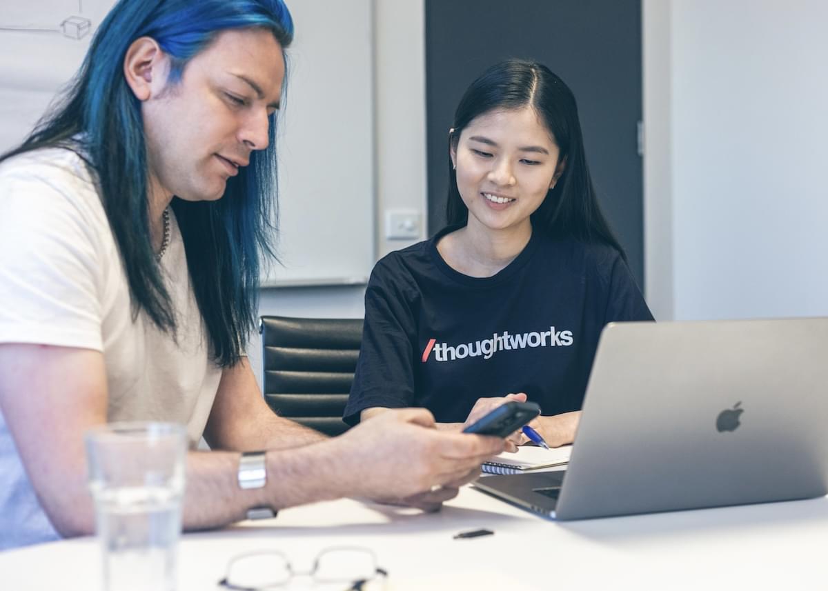 A man looks at a mobile phone as he sits next to a woman in a Thoughtworks t-shirt who is taking notes in front of a laptop