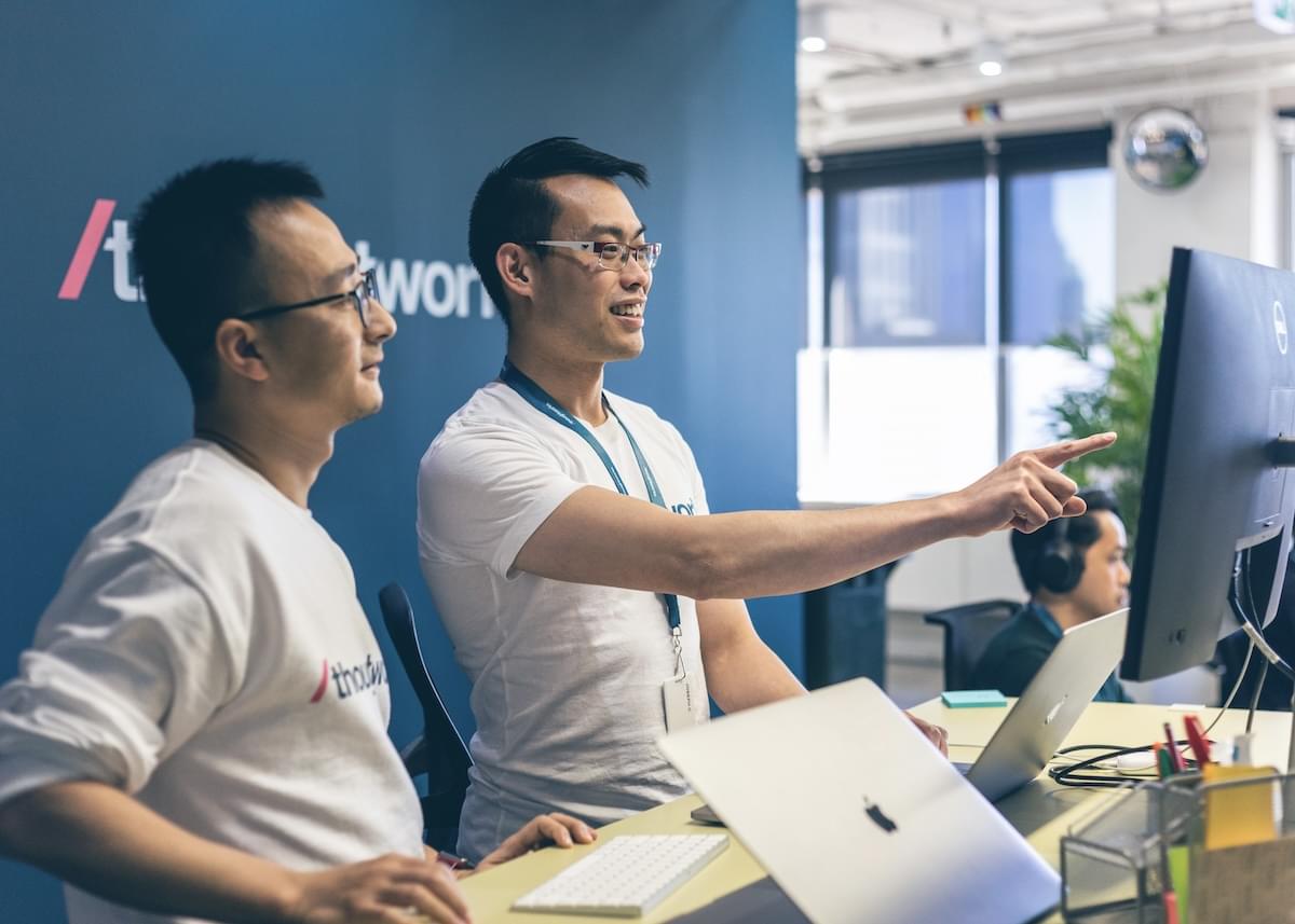 Two young men in an office look at a computer in front of a wall displaying a large Thoughtworks logo