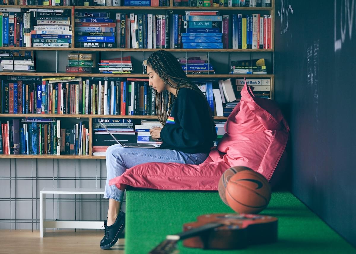 A young woman works on a laptop in front of a bookshelf filled with computer books