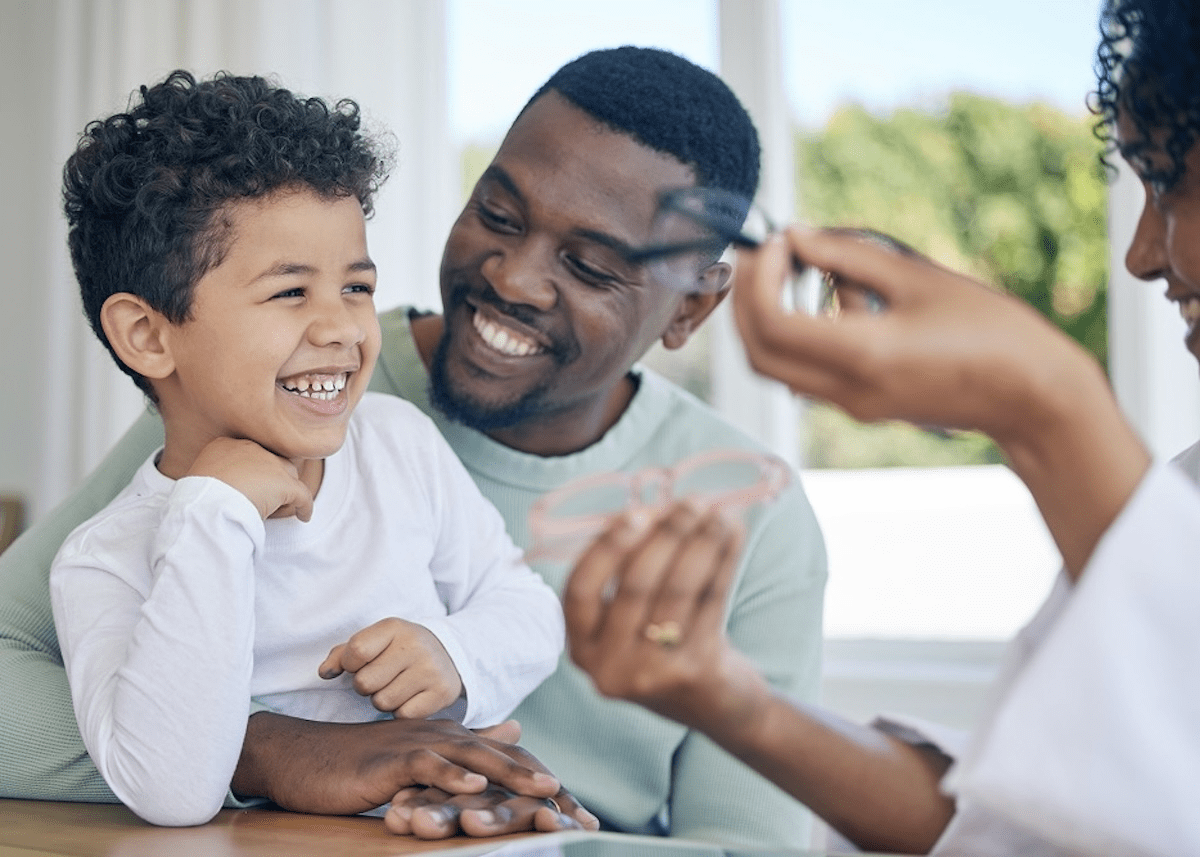 A father encourages his young son to choose a pair of glasses at the optometrist as a woman holds up a pink pair of frames and a black pair of frames