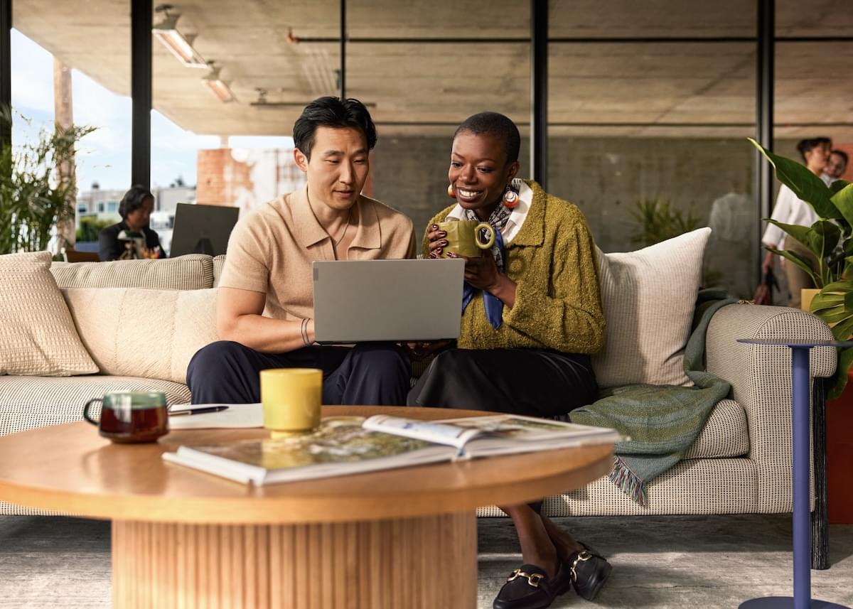 A man and a woman sit on a comfortable couch in a modern, airy office and look at a laptop together
