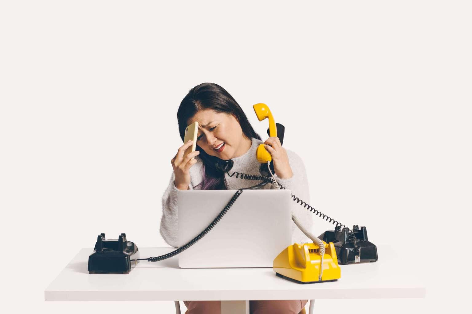 Woman working at a desk with a laptop and multiple phones that she is trying to answer all at the same time