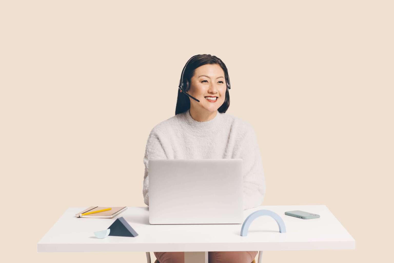 Customer service worker wearing a phone headset sitting at a desk with a laptop open and smiling