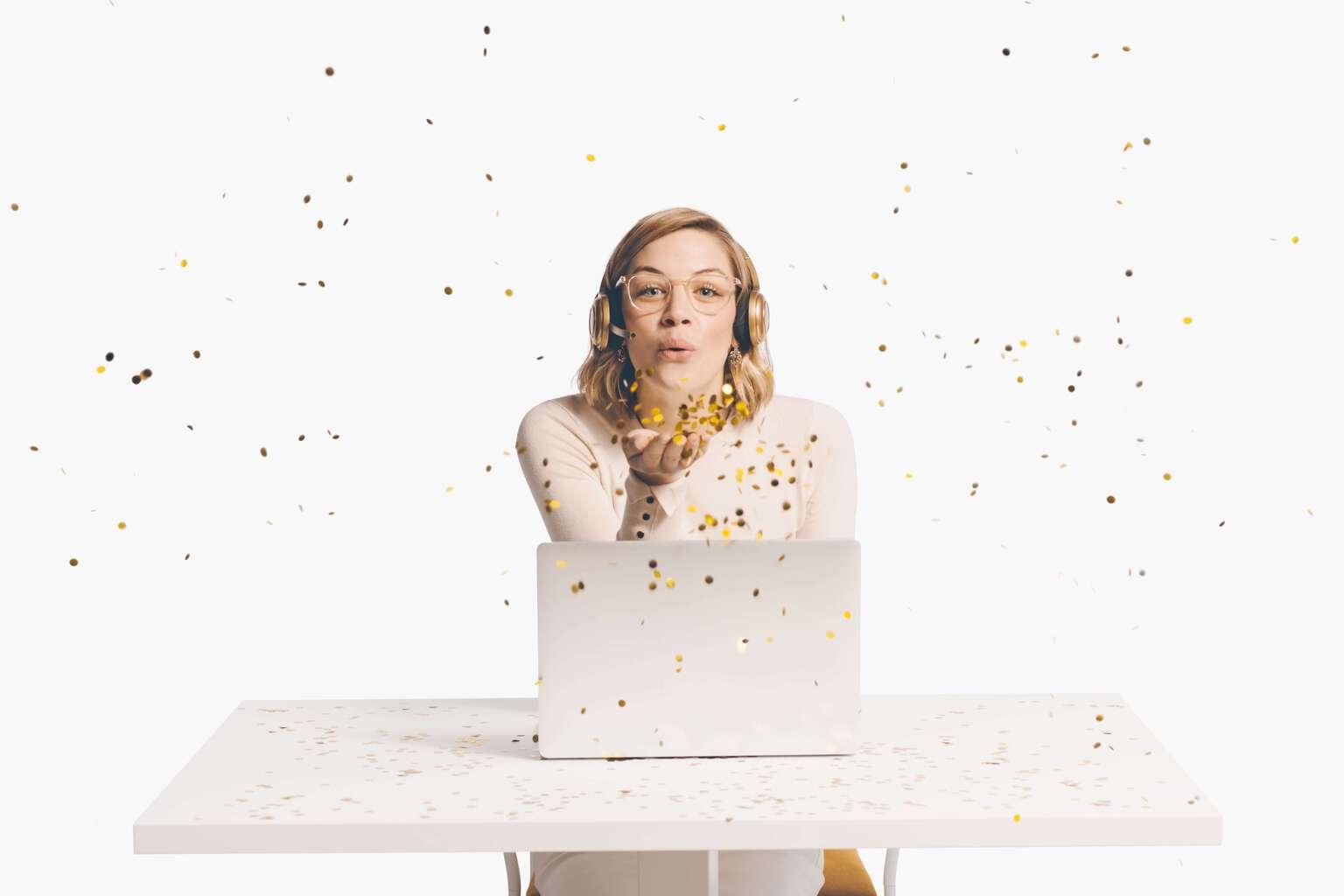 Woman wearing a customer service headset sitting at a desk blowing confetti toward the viewer playfully