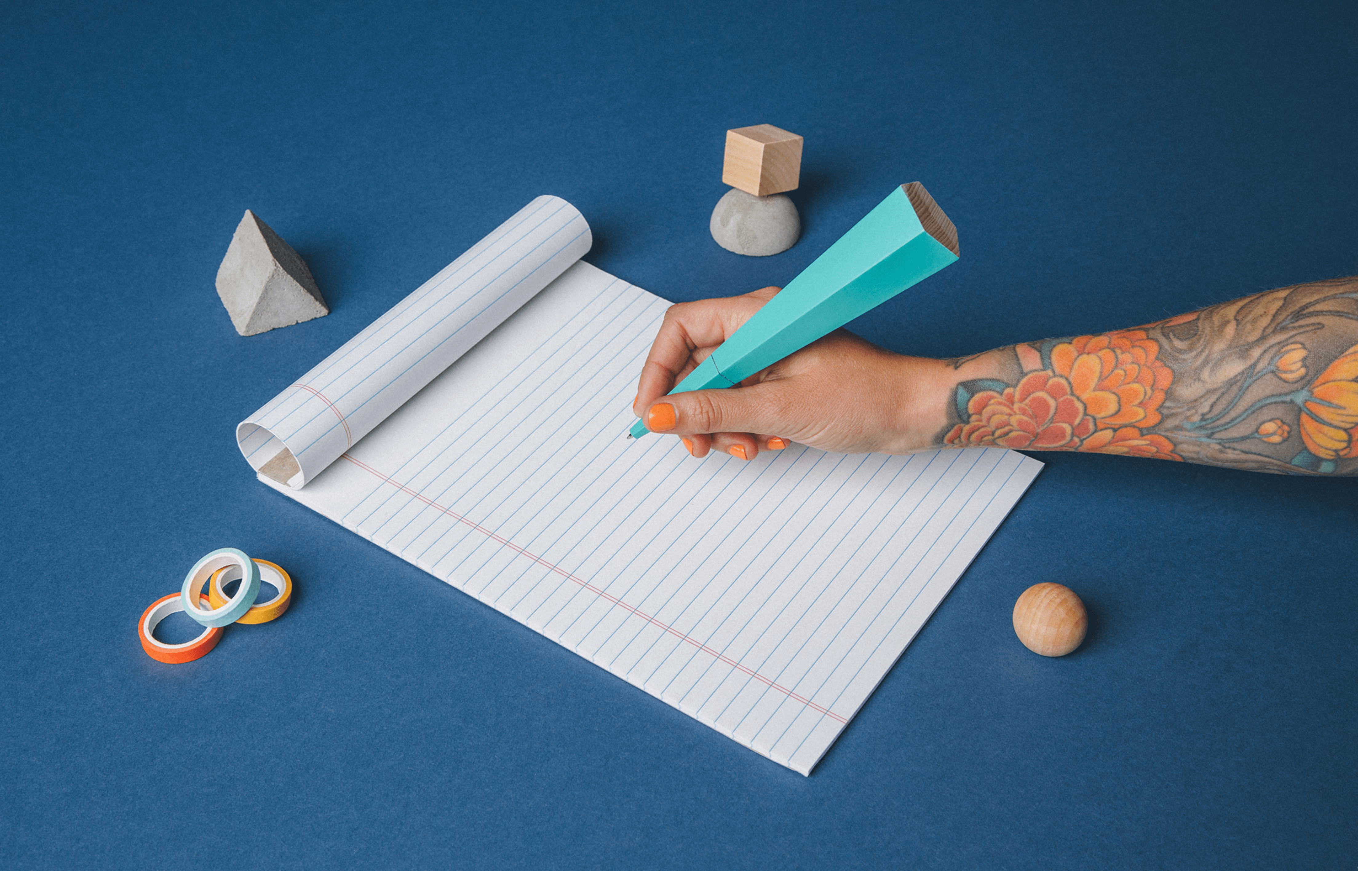 Person writing on a notepad with a uniquely shaped pen and various shapes on the table around the notebook