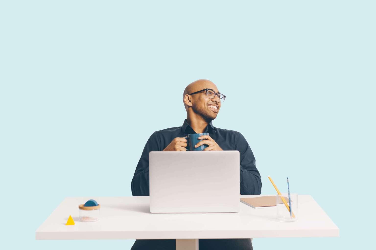 Man sitting at a desk with laptop open holding a coffee mug and smiling